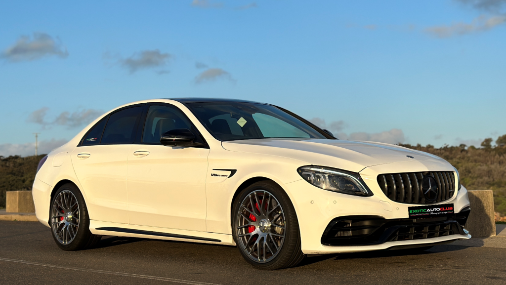 A white Mercedes-Benz C63s sports sedan is parked on a paved area with a backdrop of blue sky and some clouds. The car features a sleek design with tinted windows, red brake calipers, and a prominent front grille. The AMG logo is visible on the front and side.