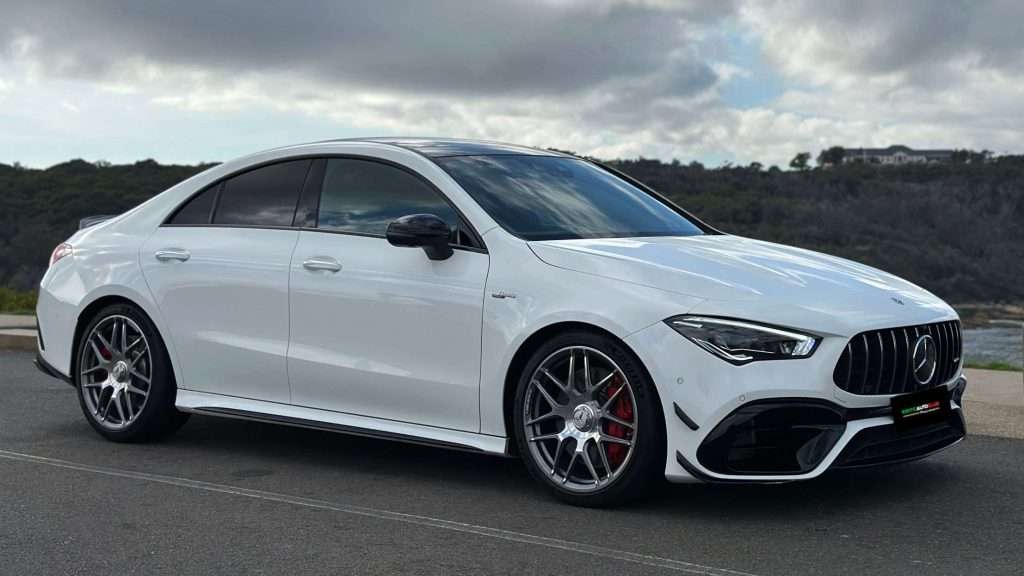 A sleek, white Mercedes-Benz CLA45s performance sedan with tinted windows and sporty red brake calipers is parked on a paved area. The background features a cloudy sky and greenery. The car's modern design includes a black grille and low-profile tires on alloy wheels.