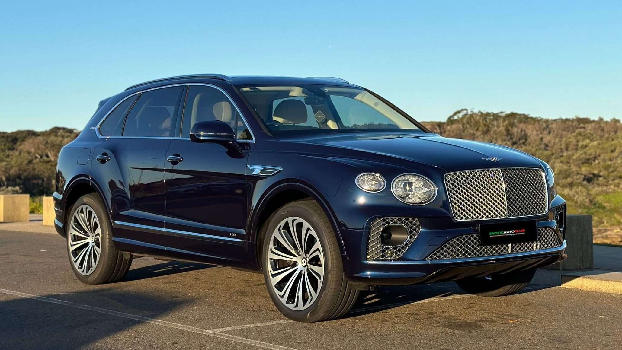 A dark blue Bentley Bentayga SUV parked outdoors on a sunny day. The car has a distinctive large front grille, sleek design, and alloy wheels. The background features greenery and clear blue sky.