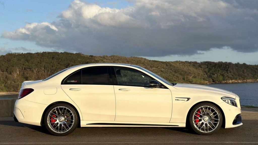 A sleek white Mercedes-Benz C63s sedan is parked on a road with lush greenery and a body of water in the background. The AMG car features silver alloy wheels, red brake calipers, and dark-tinted windows. The sky above is partly cloudy.