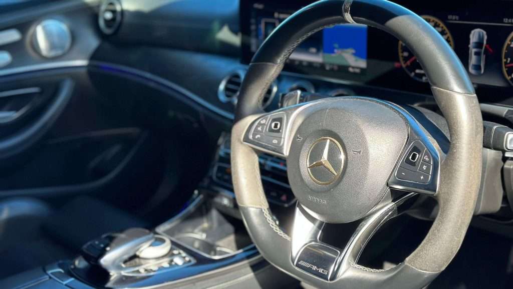 Close-up of a Mercedes-Benz E63s interior focusing on the AMG steering wheel with the Mercedes logo. The dashboard and control panel are visible in the background with a map displayed on the navigation screen. The interior features a sleek black design.