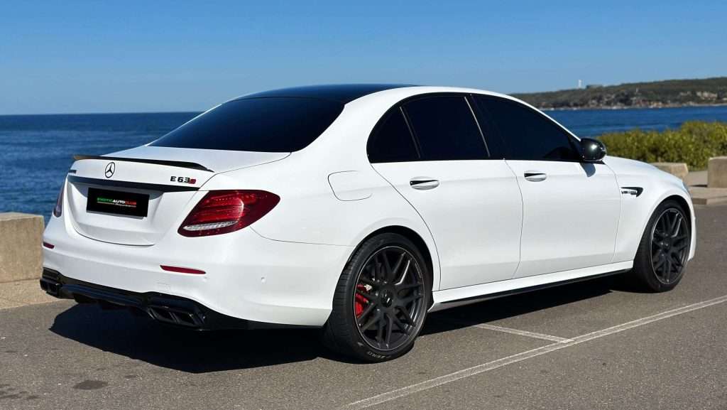 A white Mercedes-Benz E63s AMG sedan with tinted windows, black wheels, and red brake calipers is parked on a coastal road. The sea and a clear blue sky form the perfect backdrop.