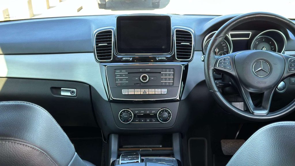 Interior view of a Mercedes-Benz GLE 250d's front cabin with a focus on the dashboard and steering wheel. The dashboard features a large touchscreen, various controls, and air vents. The steering wheel has multiple buttons and a prominent car logo in the center.