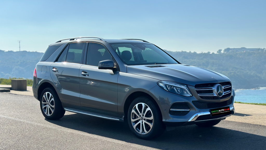 A sleek, dark gray Mercedes-Benz GLE 250d SUV is parked on a road in front of a scenic landscape with hills and clear blue sky in the background. The vehicle has a distinctive front grille with the Mercedes-Benz emblem and stylish alloy wheels.