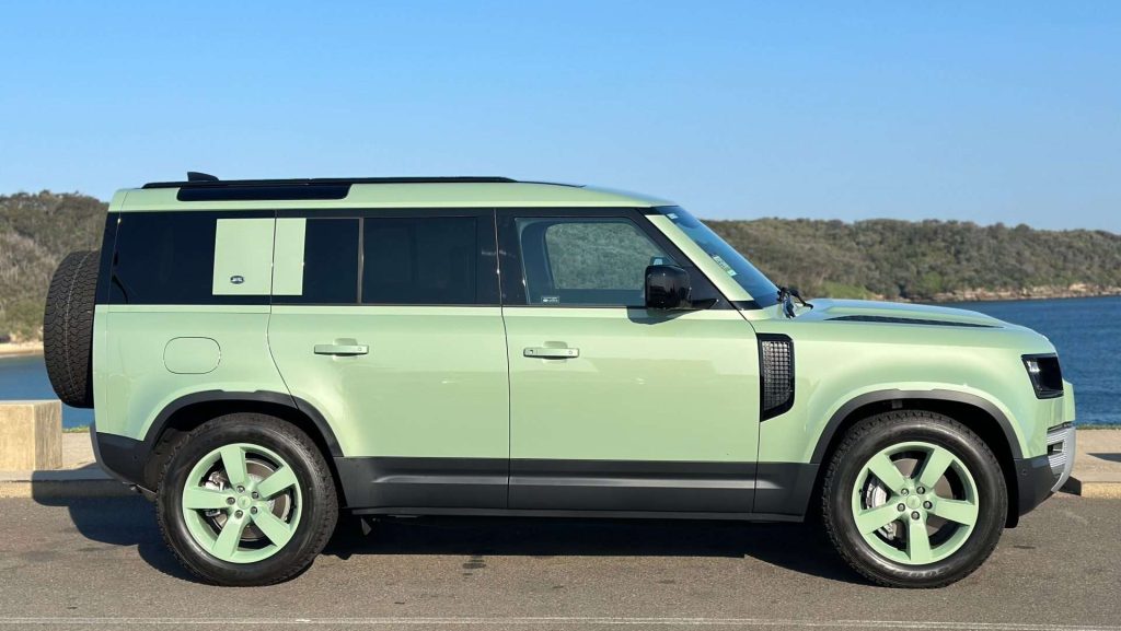 Side view of a mint green Special Edition Land Rover Defender parked near a coastal area with a body of water and green hills in the background. The SUV has a sleek, modern design with tinted windows and matching mint green wheels. The sky above is clear and blue.