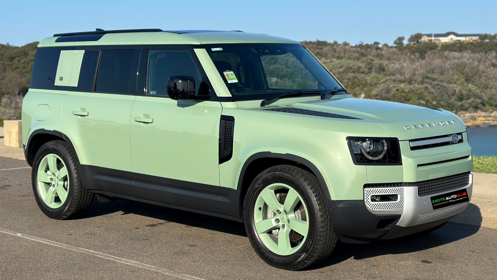 A mint green Land Rover Defender 7th Edition SUV parked outdoors against a scenic backdrop. The vehicle features black accents on the roof and door trims, silver details on the front grille, and matching mint green wheels. The surroundings include trees and hills.