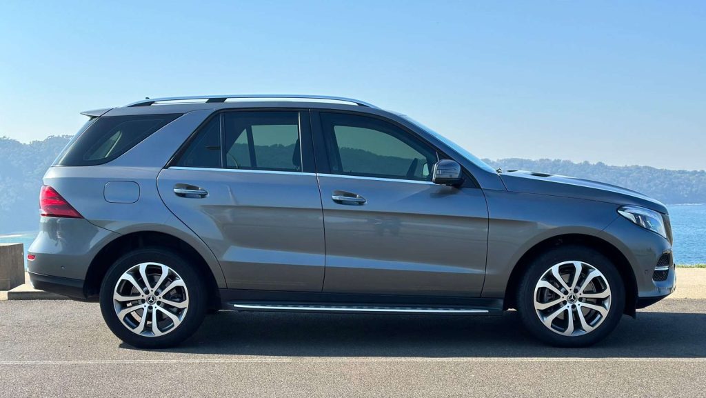 A silver Mercedes-Benz GLE 250d is parked on a paved surface with the ocean and distant mountains in the background. The vehicle is viewed from the side, showcasing its sleek design and modern curves against a clear blue sky.