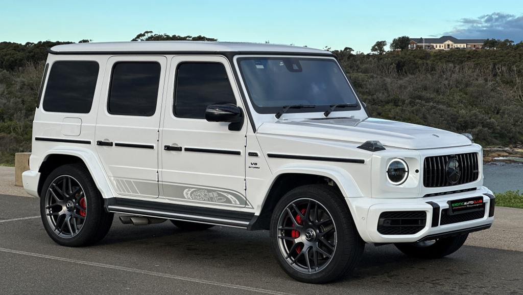 A white Mercedes-Benz G63 AMG is parked on a paved road near a body of water and greenery. The vehicle has tinted windows, black and silver rims, and red brake calipers. Celebrating its 55th Anniversary, the weather is clear with a blue sky visible in the background.