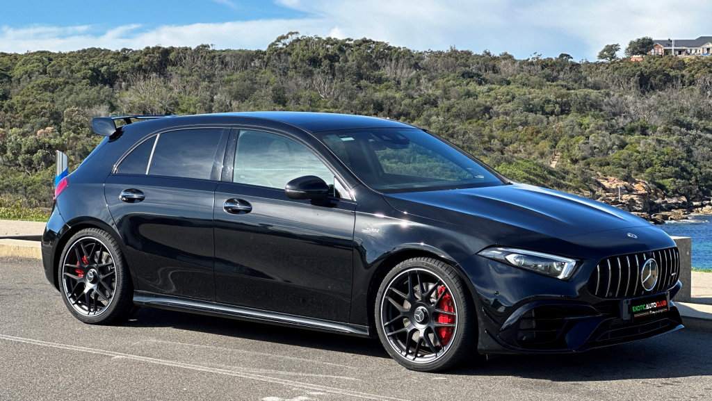A sleek black Mercedes-Benz A45s is parked on a scenic coastal road with lush greenery and a blue sky in the background. The car features red brake calipers, stylish alloy wheels, and a rear spoiler, emphasizing its sporty design.