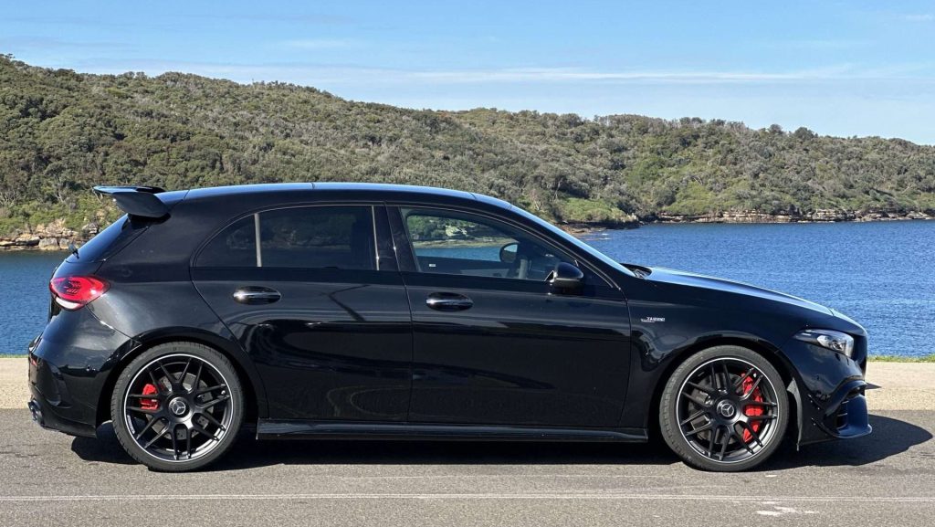 A black Mercedes-Benz A45s with tinted windows and red brake calipers is parked on a road adjacent to a body of water, with green hills and a blue sky in the background, epitomizing the essence of a luxury car.