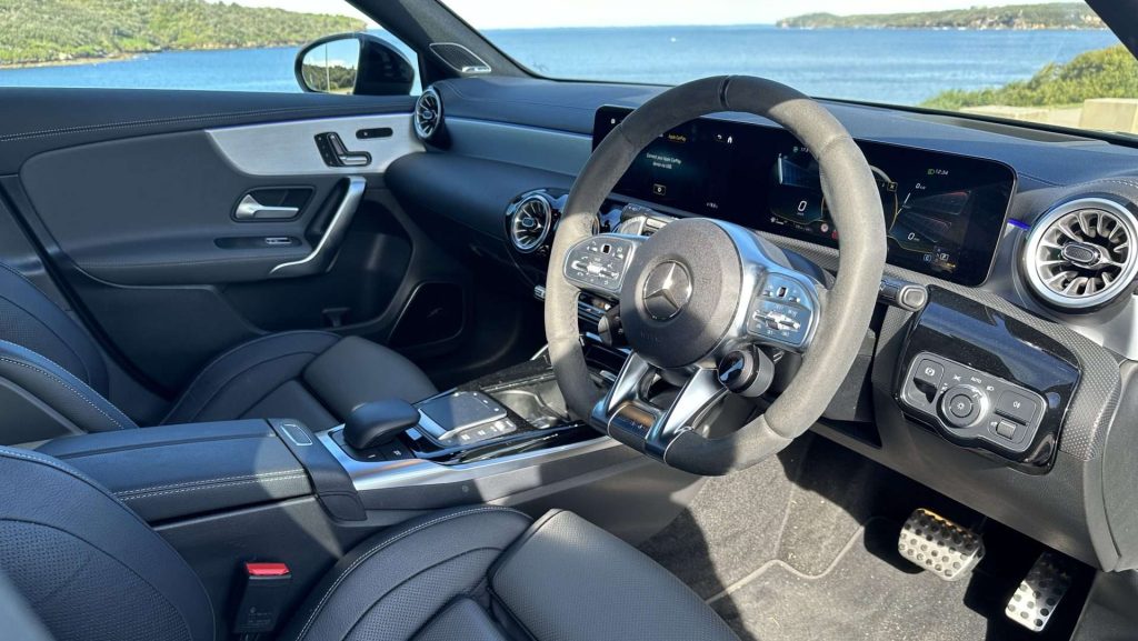 Interior view of the modern Mercedes-Benz A45s featuring a sleek dashboard, a multifunction steering wheel adorned with the iconic logo, digital displays, and well-appointed leather seats. The window shows a scenic coastal view with blue water and distant landmasses.