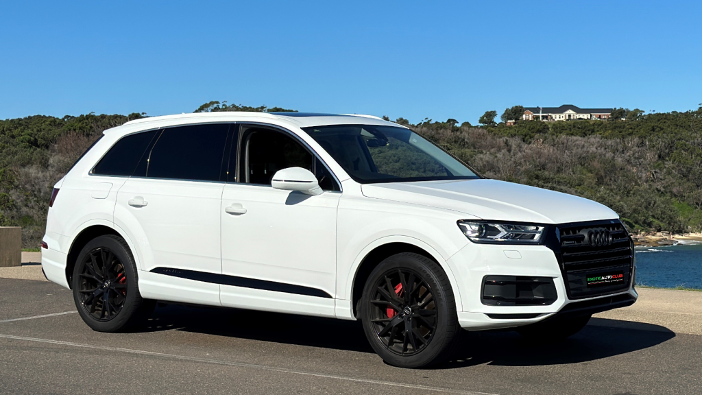 A white Audi Q7 luxury SUV is parked on a paved area next to water, with green trees and a hill in the background under a clear blue sky.