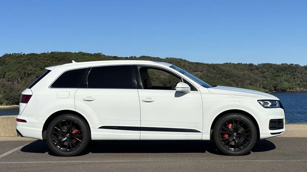 A sleek white Audi Q7 with black rims is parked on an asphalt surface. In the background, there is a body of water and a tree-covered hill under a clear blue sky. The scene suggests a serene and sunny day.