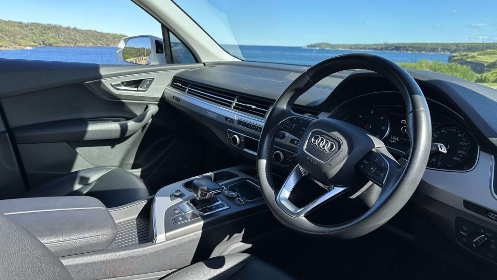 Interior view of a modern Audi Q7, showcasing the steering wheel, dashboard, and center console with various controls. The car is parked near a scenic coastal landscape with blue sky, clear waters, and green hills visible through the windows.