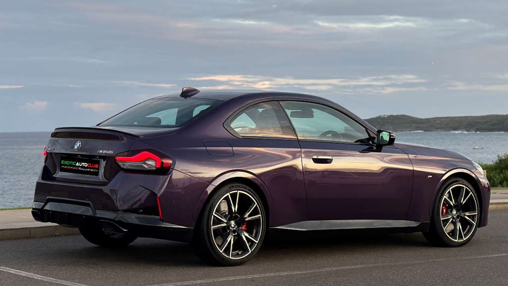 A sleek, purple BMW M240i sports coupe is parked in a scenic coastal area. The car has a modern design with sharp lines and stylish wheels. The sky is partly cloudy, and the ocean can be seen in the background, providing a serene and picturesque setting.