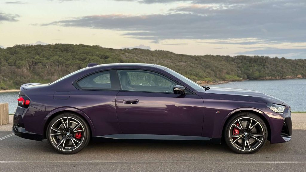 A sleek, dark purple BMW M240i is parked on a coastal road with a scenic view of the ocean and a hilly landscape in the background. The sports car features red brake calipers and stylish alloy wheels. The sky is partly cloudy with a hint of sunset light.