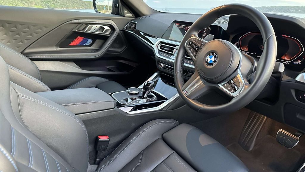 Interior of the luxurious BMW M240i, showcasing a modern dashboard, a sleek center console, and a multi-functional steering wheel proudly bearing the BMW logo. The comfortable leather seats invite relaxation while the background through the window reveals a scenic view of water and lush greenery.